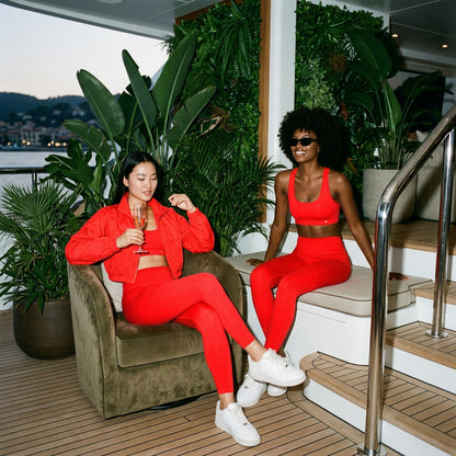 Two women in red outfits sitting on a yacht deck with plants and ocean view.